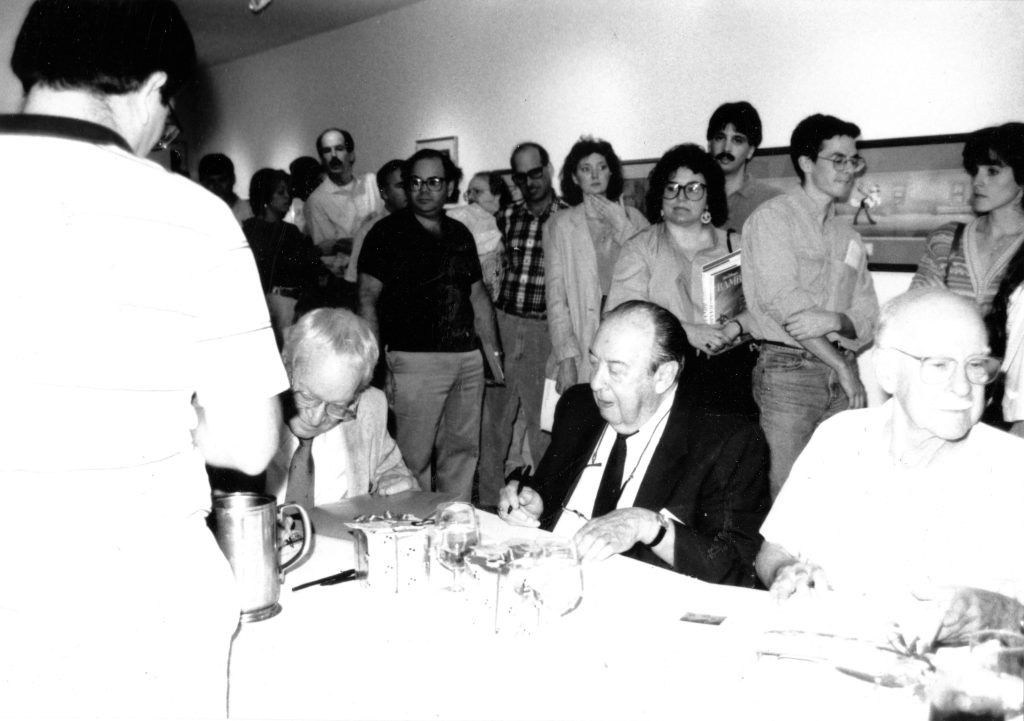 Three men sit at a long table signing books while a group of people stands behind them in a gallery setting.